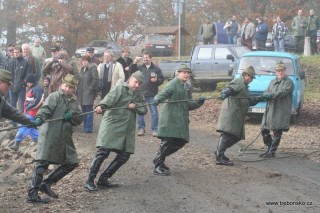 Pohled na výlov Bošileckého rybníka. Na fotografii pěšáci táhnou rozprostřenou síť ke kádišti. Práci rybářů sledují z hráze rybníka desítky návštěvníků.